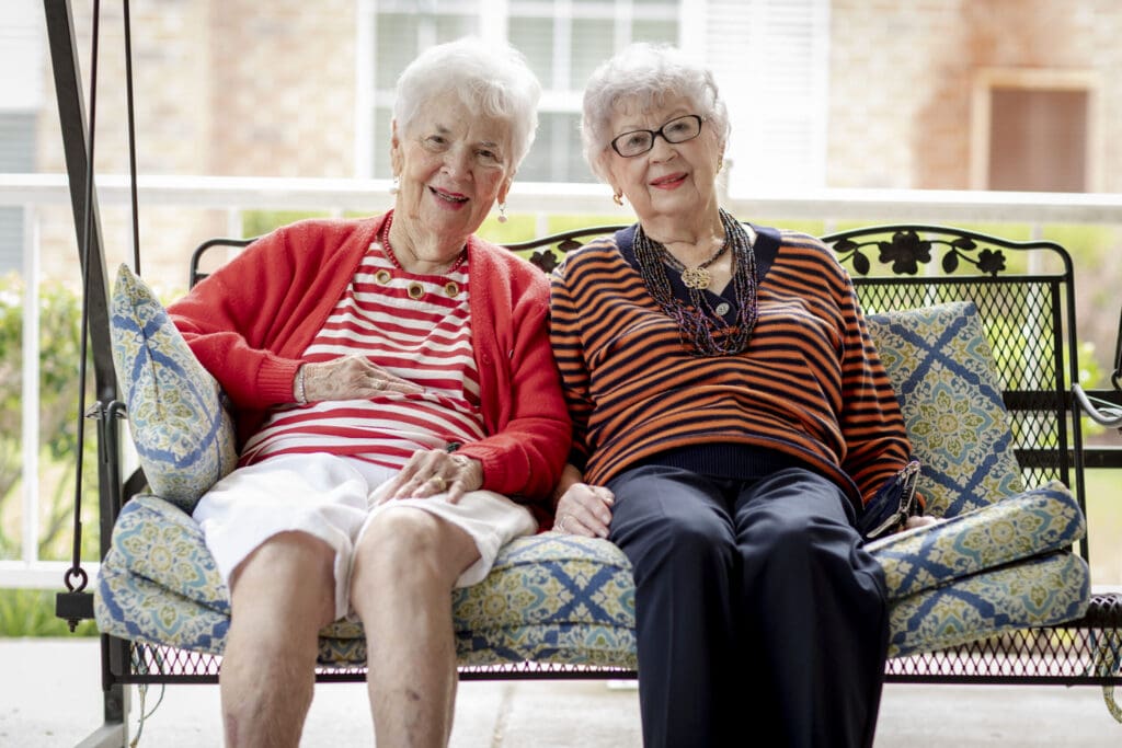 Two joyful elderly women sit together on a swing, enjoying a sunny day in a serene outdoor setting.