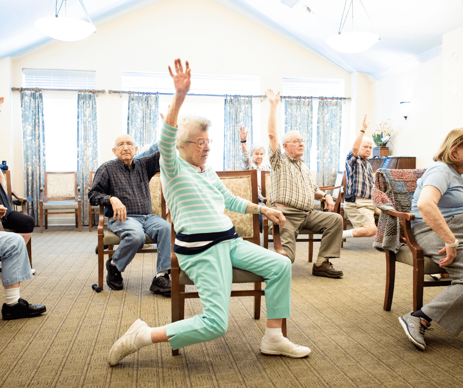 Residents engage in seated exercise, joyfully raising arms in a sunlit activity room, promoting wellness and community.