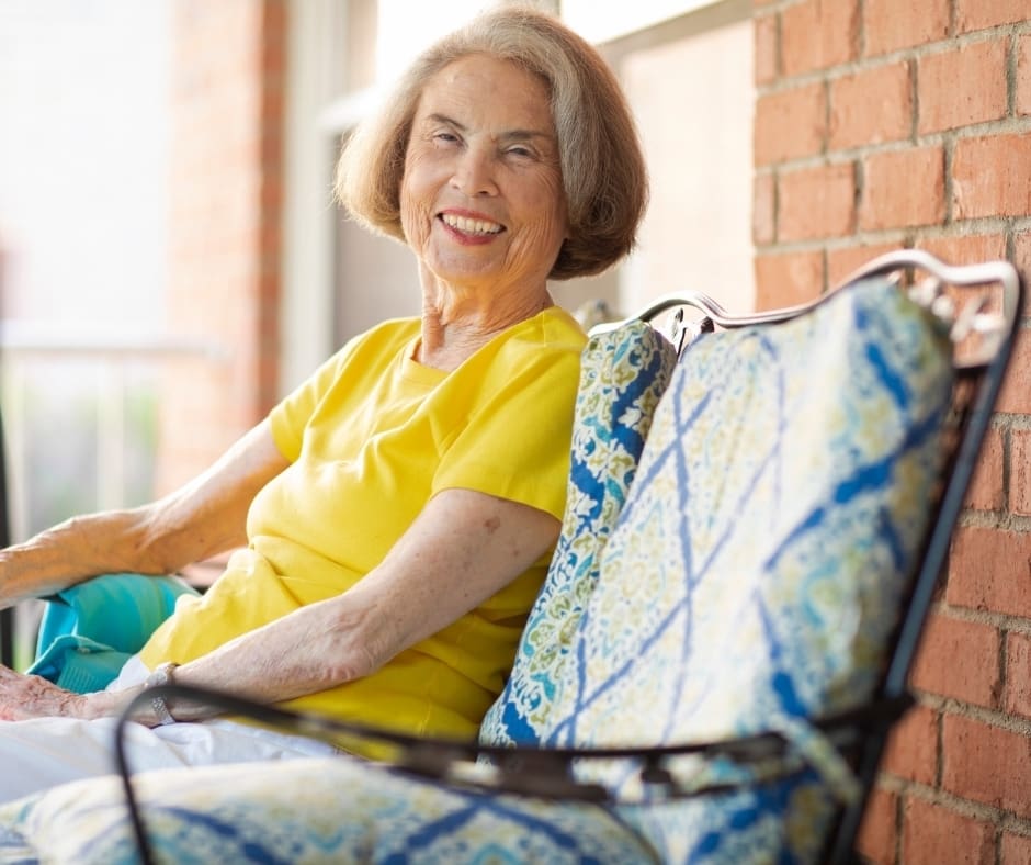Smiling senior woman in a yellow shirt sits on a vibrant patterned chair, enjoying a sunny moment on a porch.