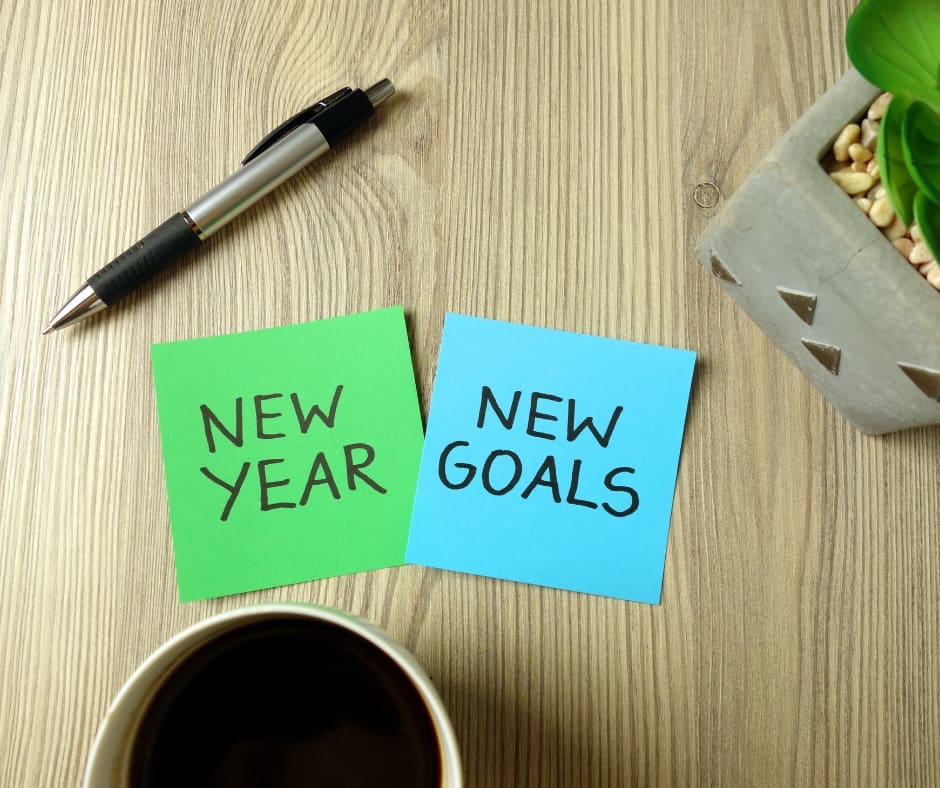 Sticky notes reading New Year and New Goals beside a pen, coffee cup, and a potted plant on a wooden table.