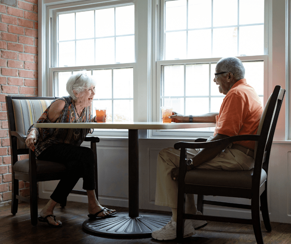 Seniors enjoying a lively conversation over iced tea at a bright, welcoming dining space with large windows.