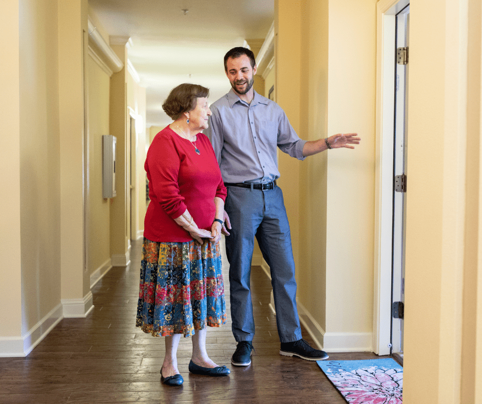 A staff member warmly engages with a resident in a bright, welcoming hallway of a senior living community.