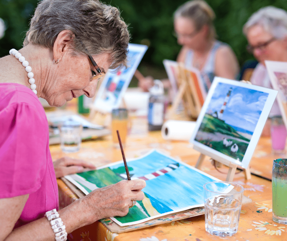 Senior woman painting at an outdoor art class, surrounded by peers, showcasing creativity and vibrant social interaction.