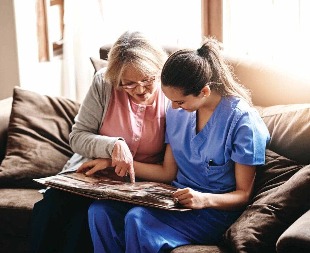 A caregiver and a senior woman share a joyful moment, looking through a photo album on a cozy sofa in a warm setting.