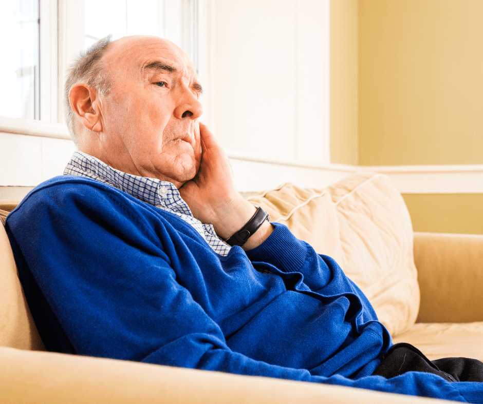 Thoughtful senior man in a blue sweater sits on a couch, gazing out a window in a bright, welcoming room.
