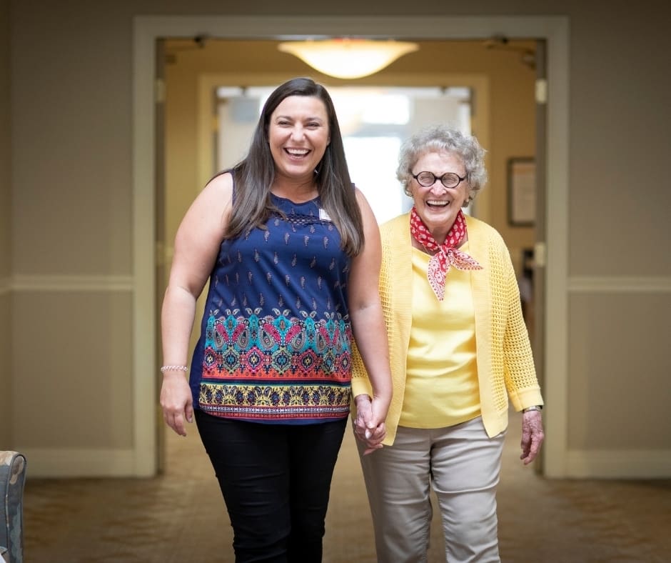 Two women, smiling and holding hands, walk joyfully down a bright, welcoming hallway in a senior living community.