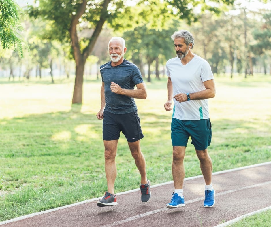 Two older men jog together on a track in a lush, green park, embodying a joyful and active lifestyle.