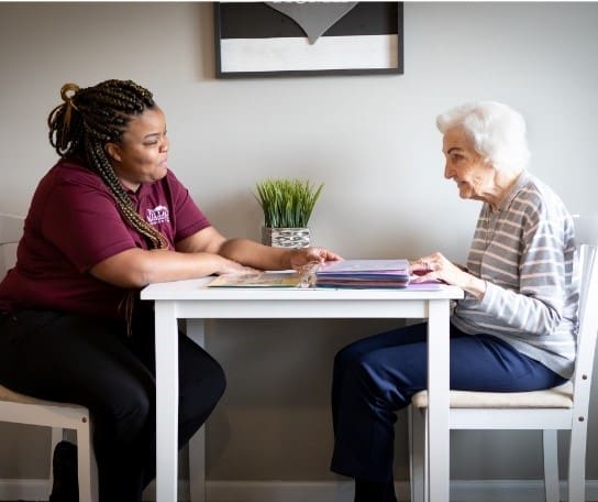 Compassionate engagement between a caregiver and resident at a bright, welcoming table in a senior living community.