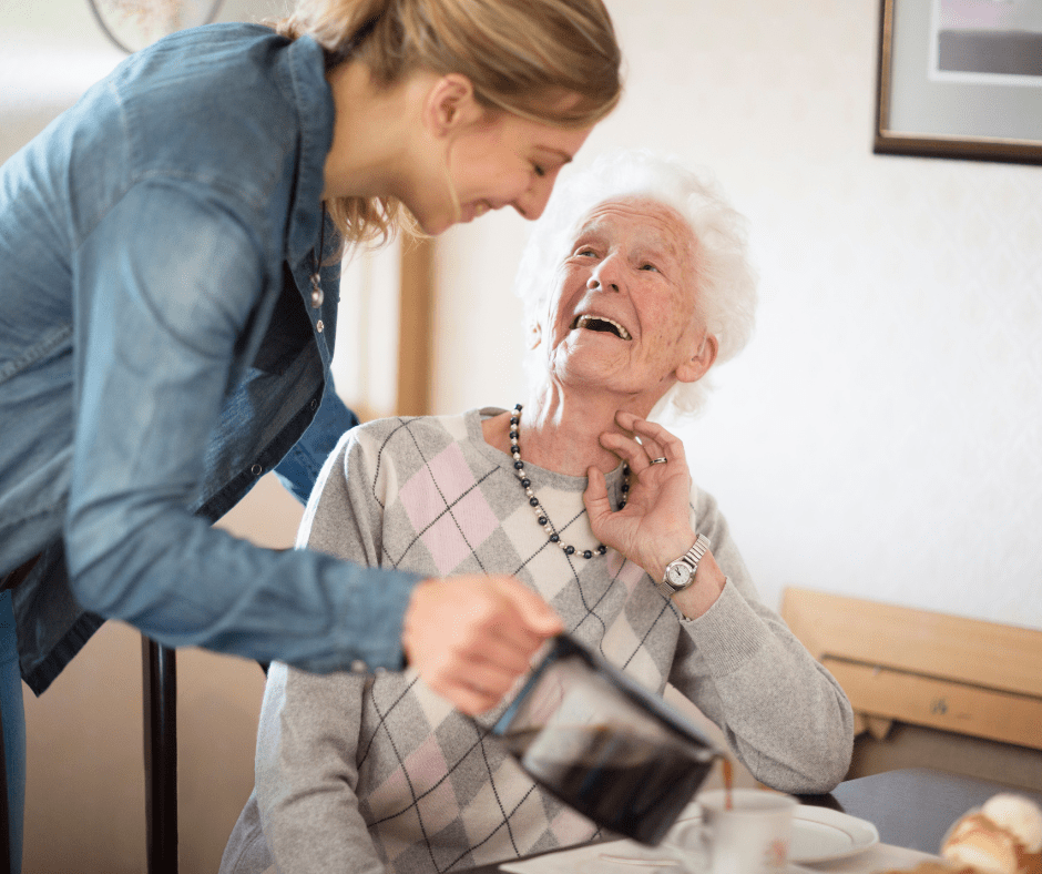 A caregiver pours coffee for a smiling senior woman in a warm, inviting dining setting, emphasizing companionship and care.