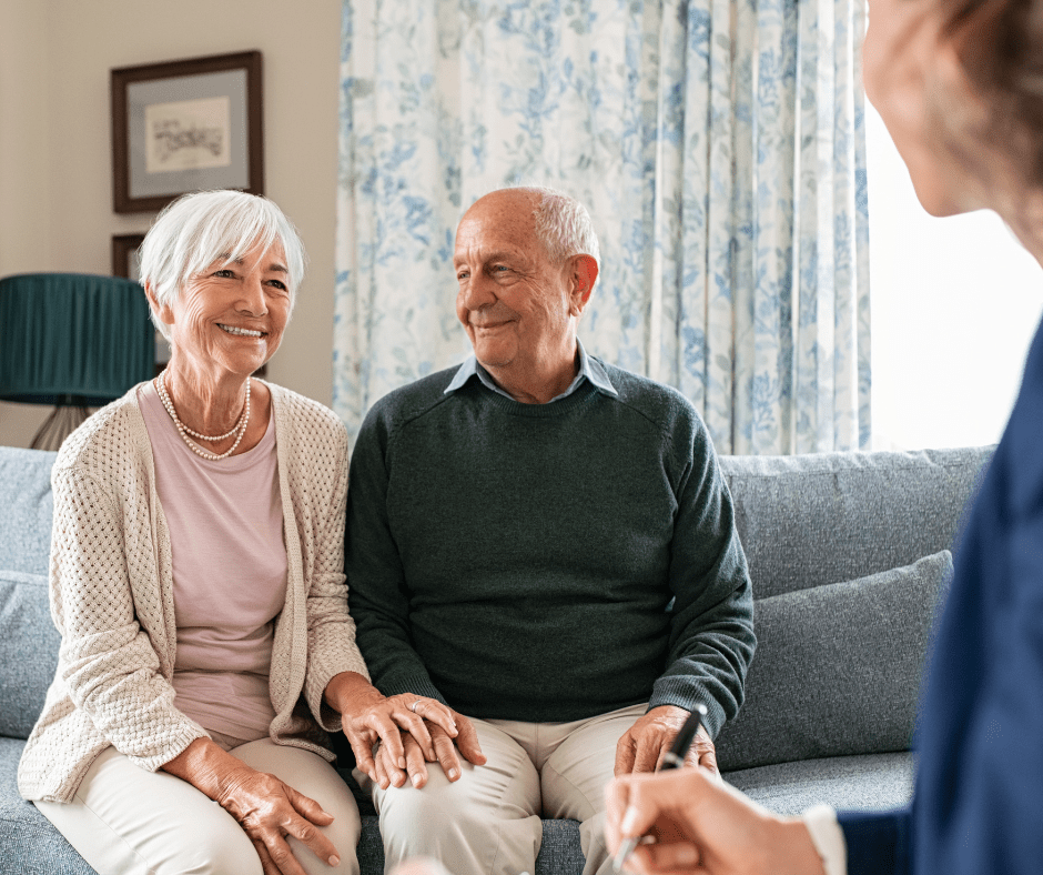 A joyful senior couple sits together on a sofa, holding hands, while engaging warmly with a caregiver.