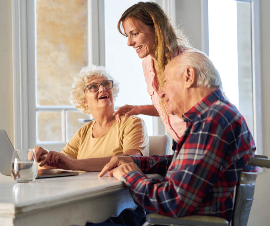 A caregiver assists an elderly couple at a bright table, fostering connection and engagement over a laptop.
