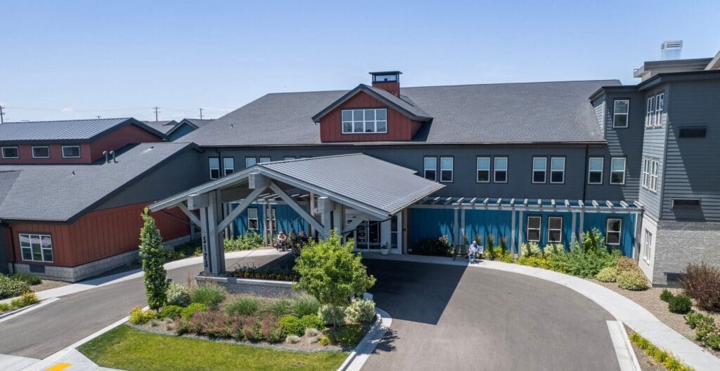 Welcoming entrance of a modern senior living community, featuring vibrant landscaping and residents enjoying outdoor seating.