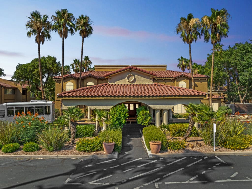Elegant senior living community entrance adorned with lush landscaping and palm trees under a clear blue sky.