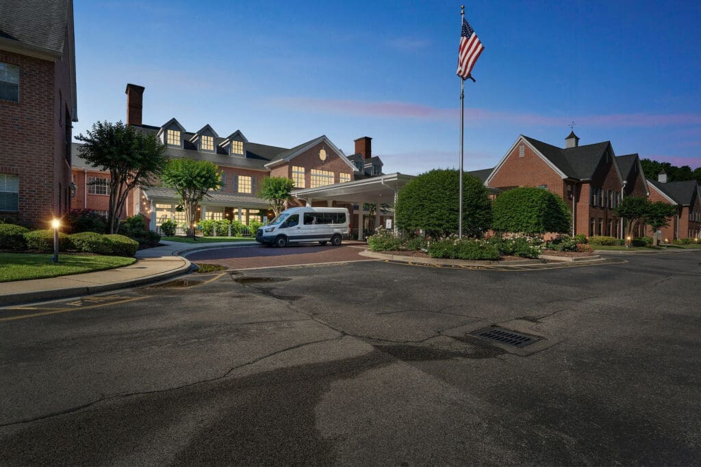 Welcoming senior living community entrance featuring lush landscaping, a shuttle van, and a flag flying proudly at dusk.