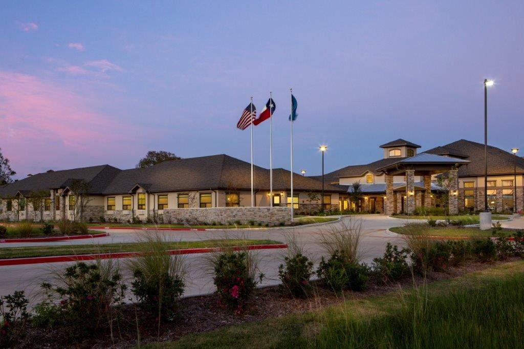 Upscale senior living community entrance with flagpoles, vibrant landscaping, and a serene twilight sky.
