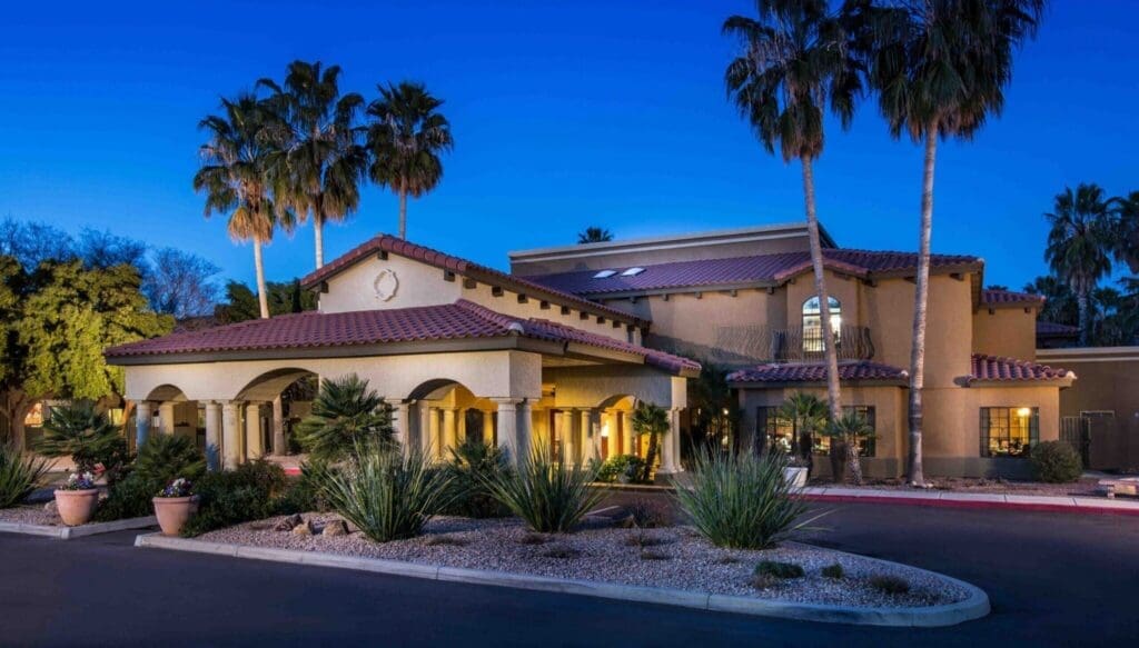 Elegant senior living community entrance with palm trees, lush landscaping, and warm evening lighting.