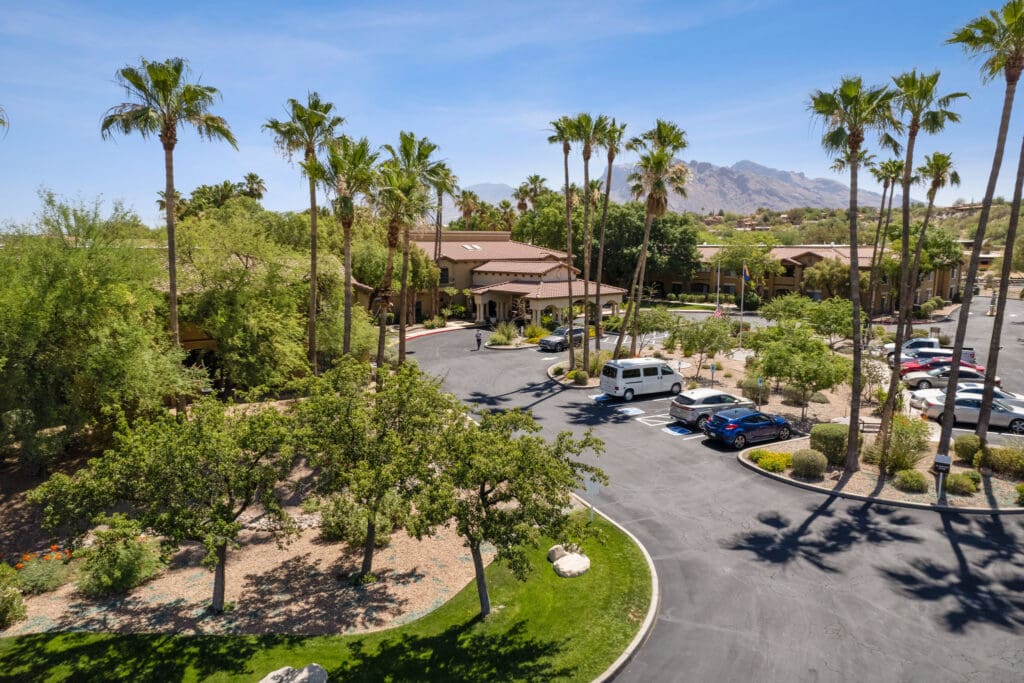 Lush landscaping surrounds a welcoming senior living community, featuring palm trees, parking lots, and distant mountains.