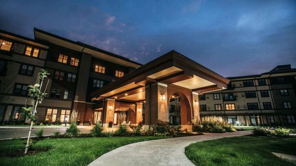 Elegant entrance of a senior living community, showcasing upscale architecture, soft lighting, and lush landscaping at twilight.