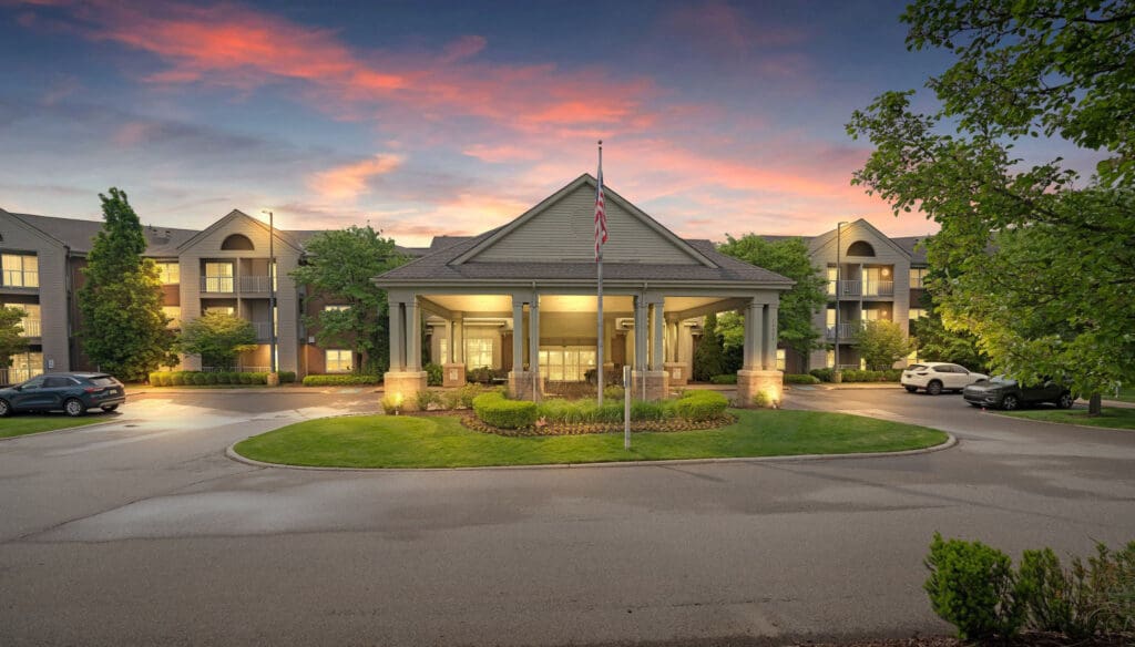 Charming entrance of a senior living community, featuring upscale architecture, lush landscaping, and a serene sunset backdrop.