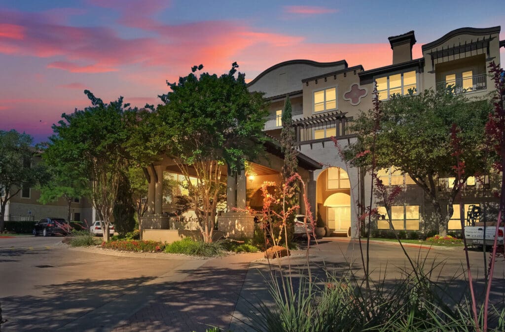 Elegant senior living community entrance at dusk, featuring lush landscaping and warm, inviting lights.