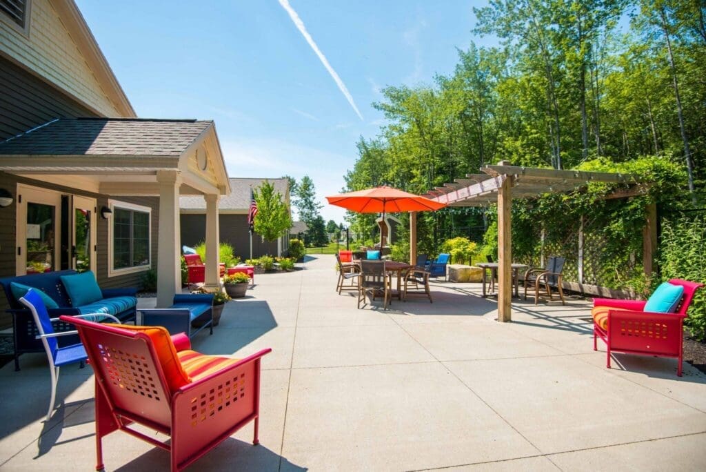 Sunny outdoor patio area with bright red, blue, and orange chairs, a red umbrella, and string lights, beside a beige house and tall green trees in the background.