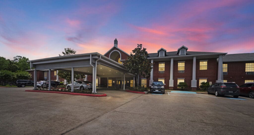 Charming senior living community entrance at dusk, featuring upscale architecture, lush landscaping, and resident parking.