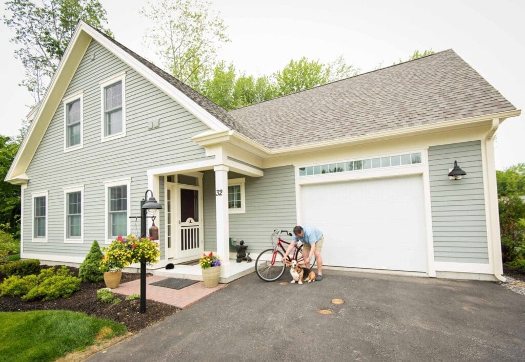 A resident interacts with two dogs near their charming grey home, featuring a bike and vibrant flower pots enhancing the entryway.
