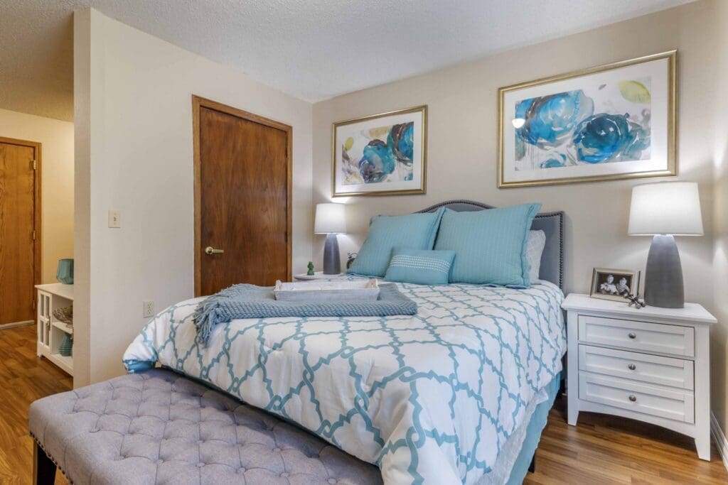 Elegant bedroom featuring a stylish bed with plush blue and white bedding, framed floral art, and warm wooden accents.