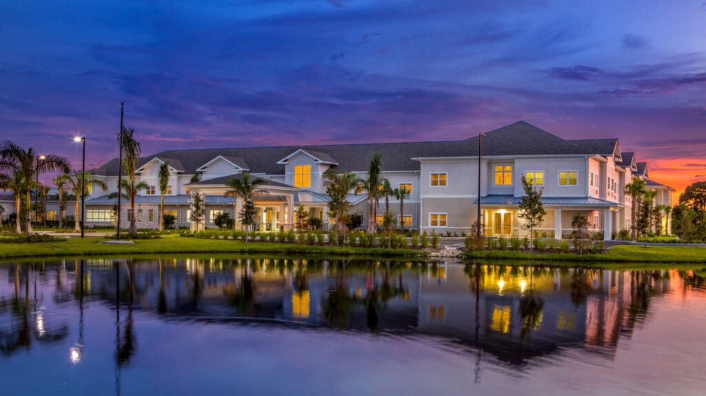 Elegant senior living community at dusk, featuring lush greenery and tranquil pond reflecting vibrant sunset colors.