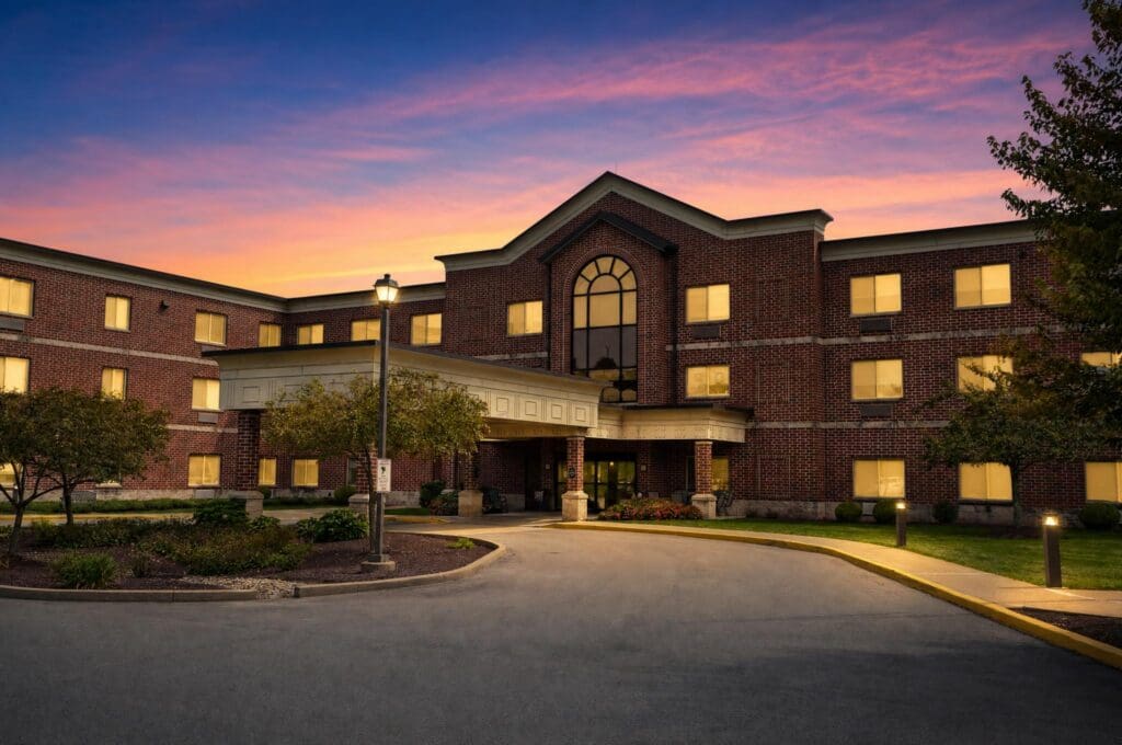 Elegant brick senior living facility at dusk, featuring warm lights, landscaped grounds, and a welcoming entrance.