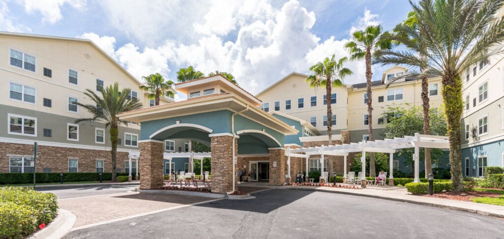 Elegant entrance of a senior living facility, featuring palm trees, stone accents, and welcoming seating areas.