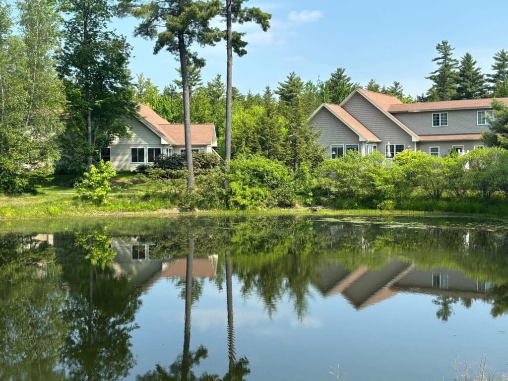 Quaint senior living residences reflect in a serene pond, surrounded by lush greenery and tall trees under a clear blue sky.