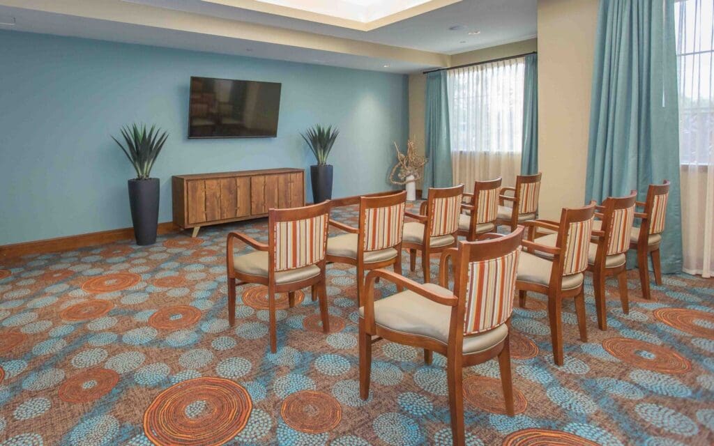Sage-Desert-Tucson-AZ-2018-June-0235-2048x1280 - Grace Management Bright and inviting community room featuring striped chairs, a large TV, and decorative plants on a vibrant patterned carpet.