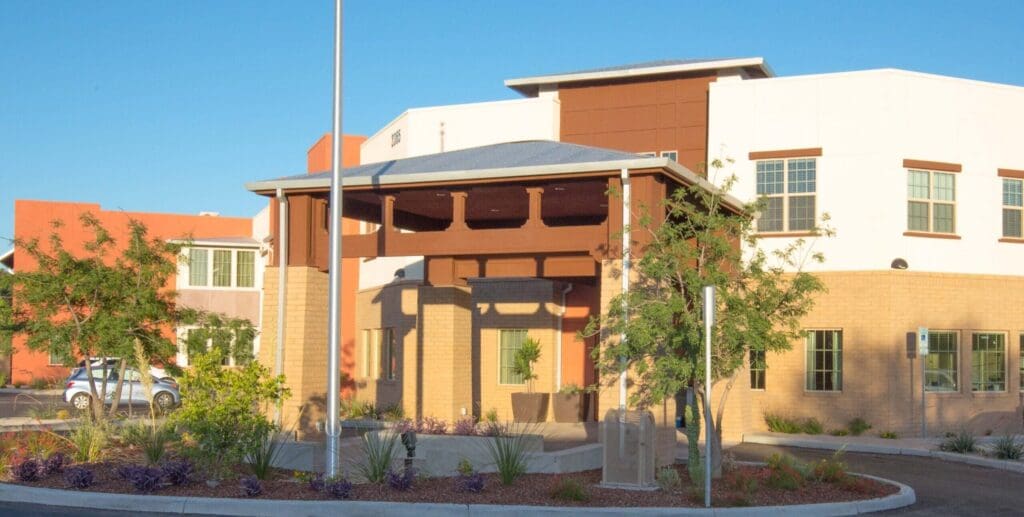Bright, modern senior living facility entrance with lush landscaping and welcoming architecture under clear blue skies.