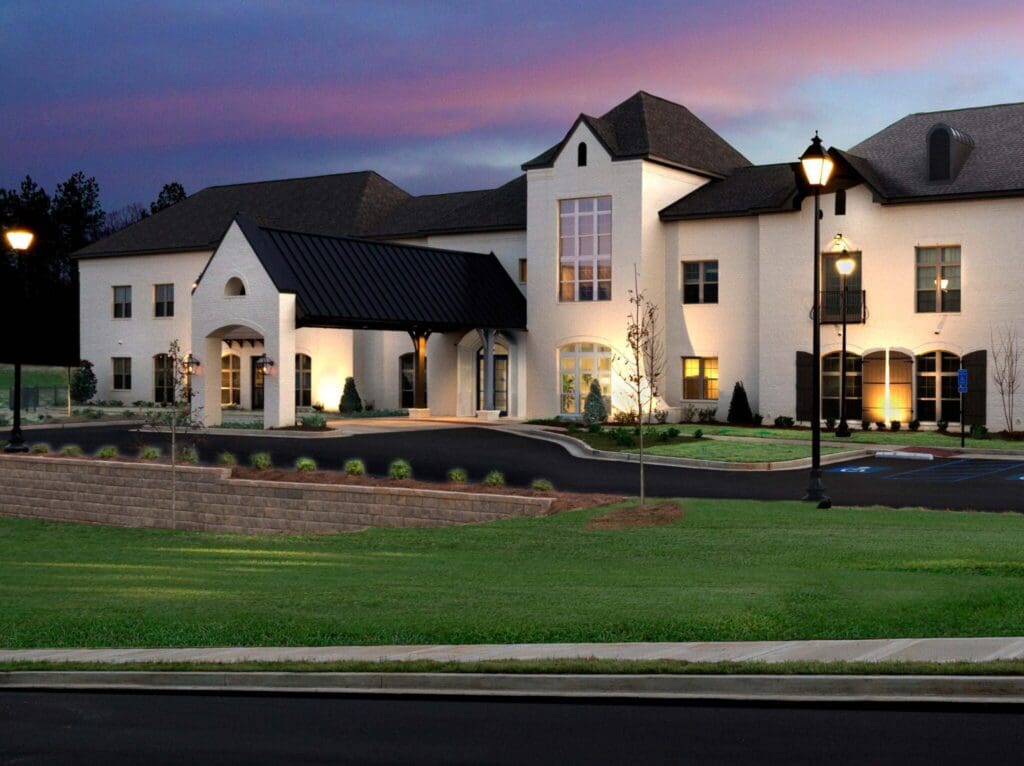 Elegantly designed senior living community featuring warm lighting, lush landscaping, and inviting entryway at dusk.
