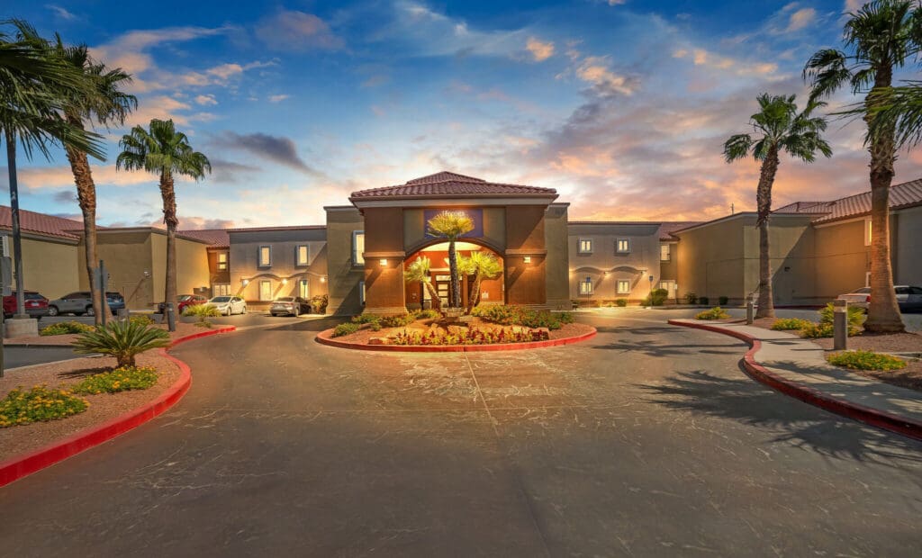 An inviting entrance of an upscale senior living community, featuring palm trees and vibrant landscaping against a sunset sky.