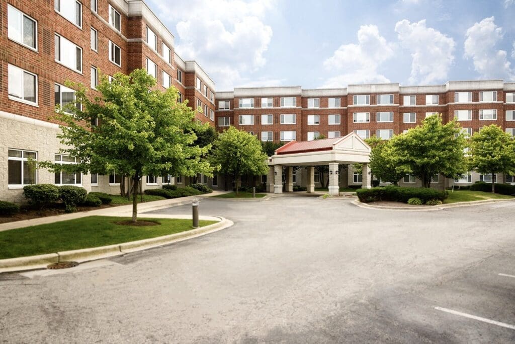 Inviting exterior of a senior living facility, featuring lush landscaping and a welcoming entrance under a bright sky.