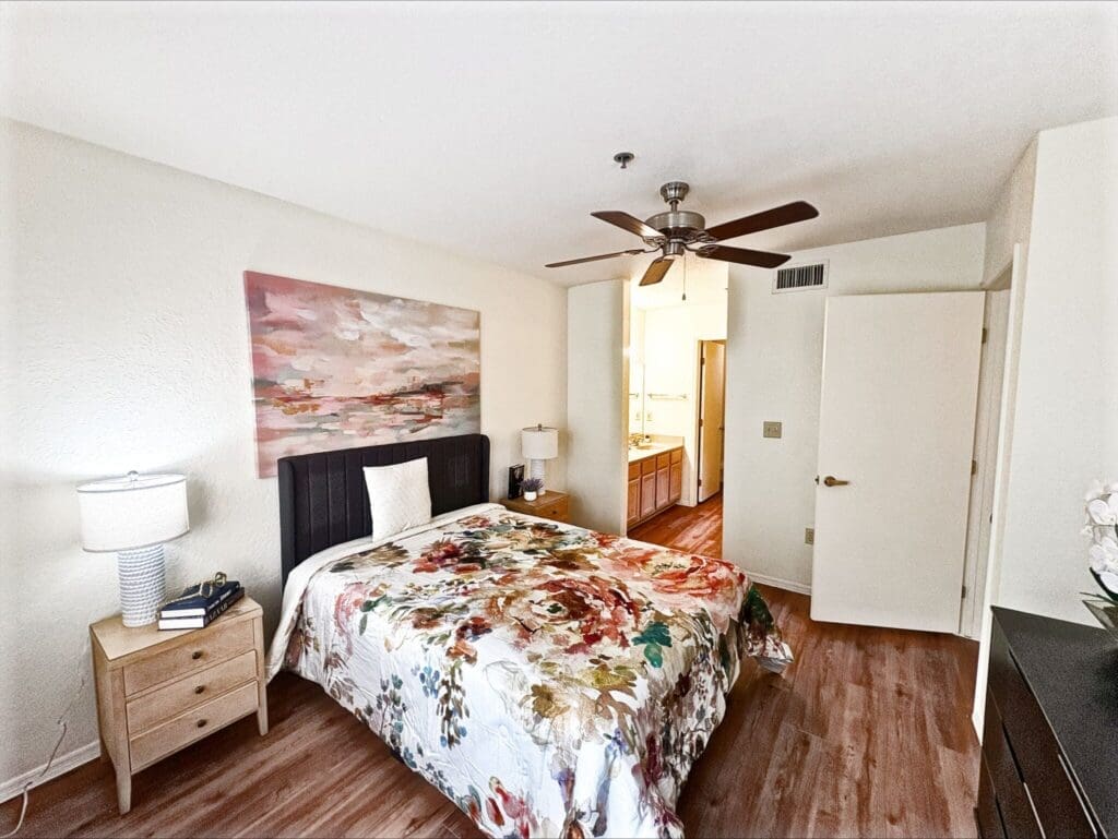 Bright, spacious bedroom with a floral quilted bedspread, modern furnishings, and an ensuite bathroom visible in the background.