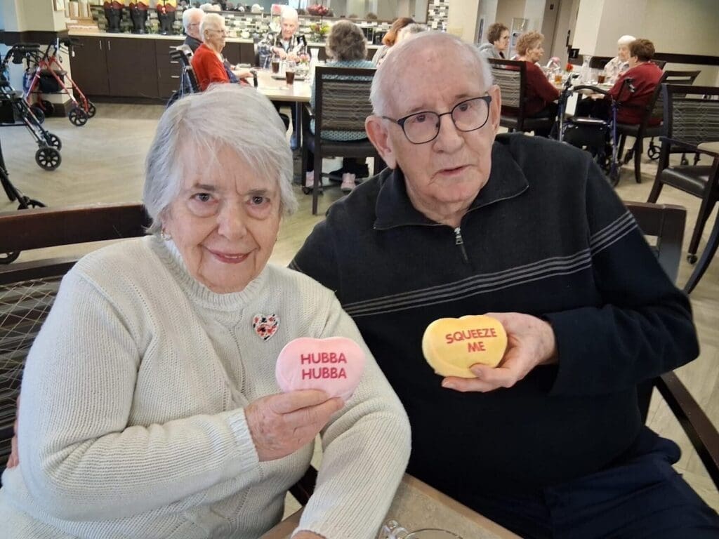 Senior couple joyfully holding heart-shaped cushions with playful messages in a vibrant dining area, surrounded by engaged residents.