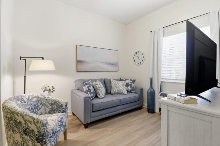 A bedroom with a small bed and blue sheets, throw pillows, a blue throw blanket, bedside table with a photo frame to the left of the bed, and a dresser with a lamp on it that sits underneath the window at Beach House, a senior living community.