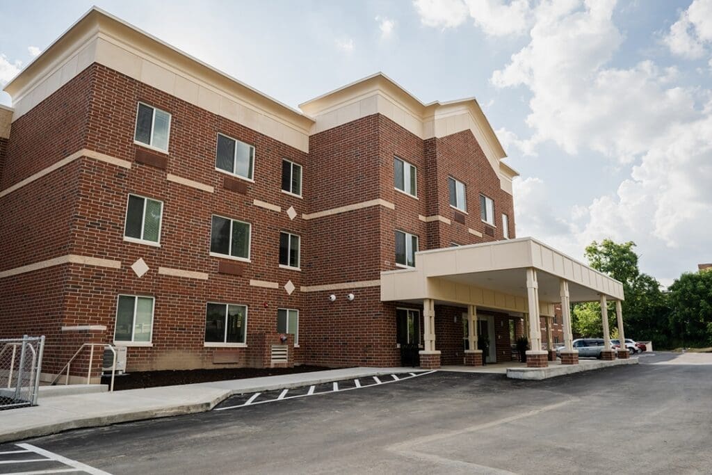 Elegant senior living facility with modern brick exterior and welcoming entrance, surrounded by lush greenery and parking area.