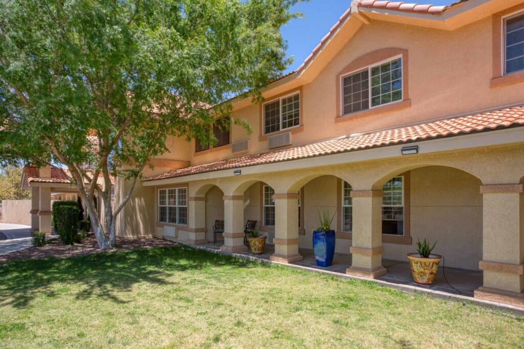 Elegant senior living community exterior featuring a welcoming porch, lush greenery, and vibrant planters under clear blue skies.