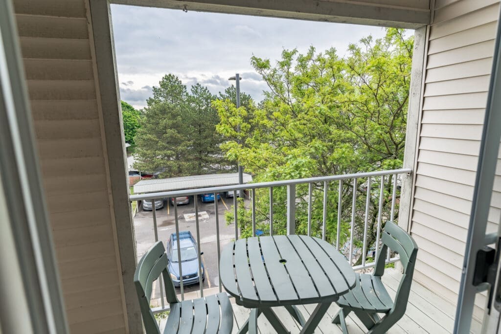 Inviting balcony with a round table and chairs, offering views of lush trees and nearby parking.
