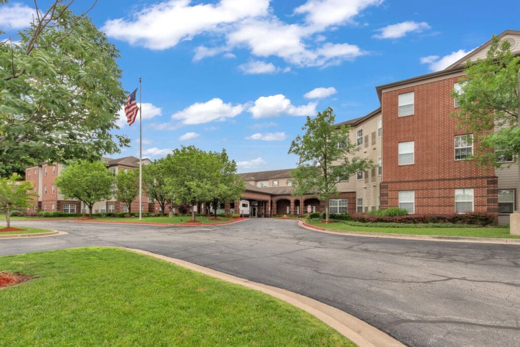 2022-05-26-HomeJab-007-1 - Grace Management Welcoming entrance of a senior living community, featuring lush greenery, a flagpole, and elegant brick architecture.