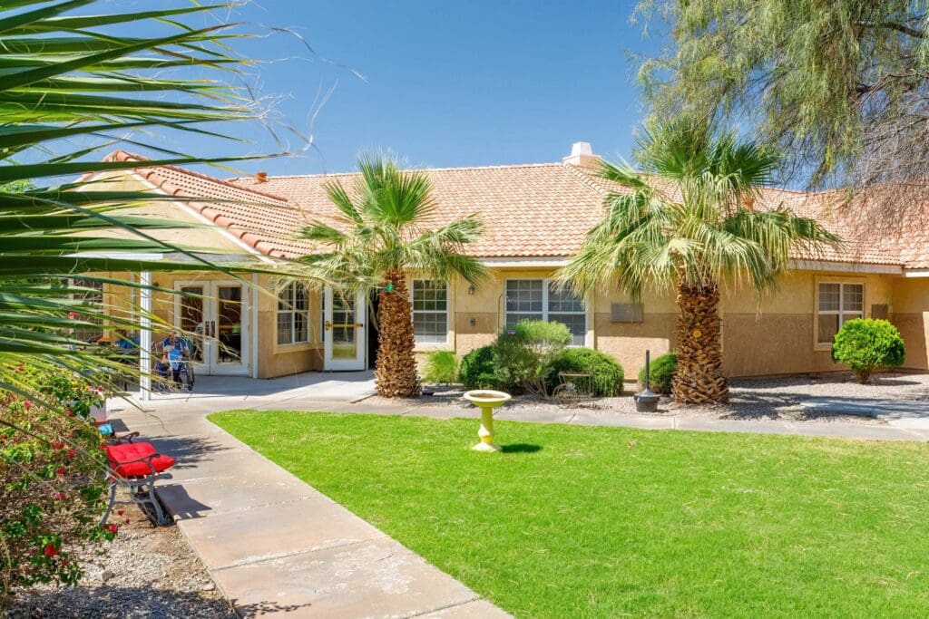 2022_LakeHavasu_EXT-Courtyard_75-scaled - Grace Management Elegant senior living community showcasing lush gardens, palm trees, and welcoming entryway under a clear blue sky.