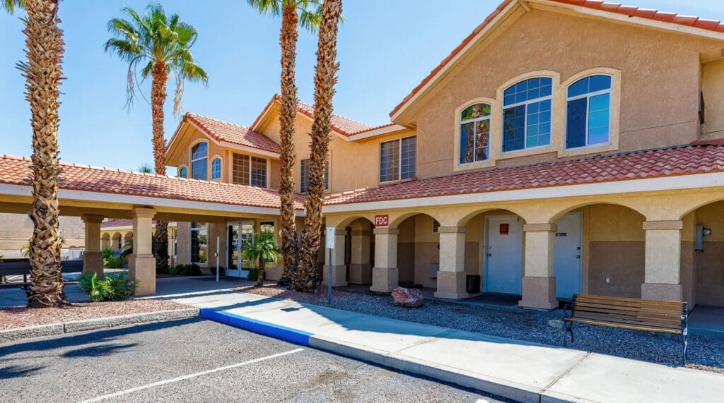 2022_LakeHavasu_EXT-Front_57-scaled - Grace Management Welcoming entrance of a senior living facility with palm trees, vibrant landscaping, and a spacious shaded seating area.