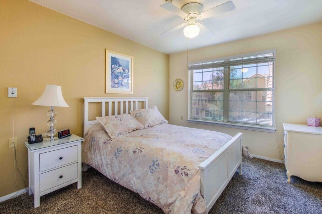 2022_LakeHavasu_INT-Model_66-scaled - Grace Management Bright, cozy bedroom featuring a white bed, floral bedding, nightstand, and window with a view of the outdoors.
