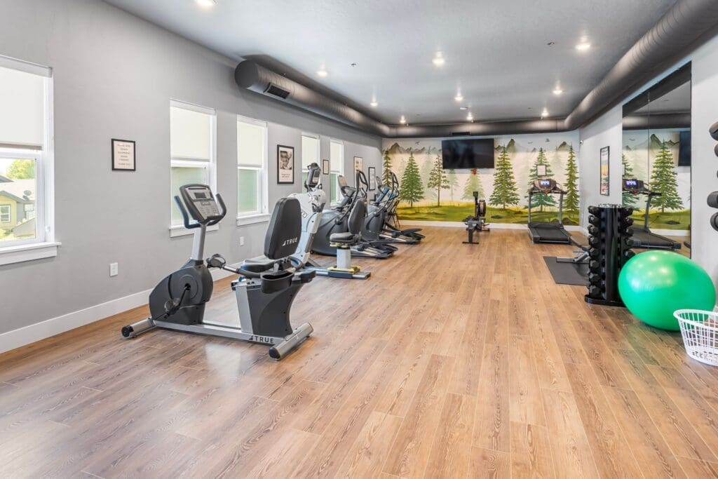 Bright fitness center featuring modern exercise equipment, large windows, and a serene nature mural enhancing the vibrant atmosphere.