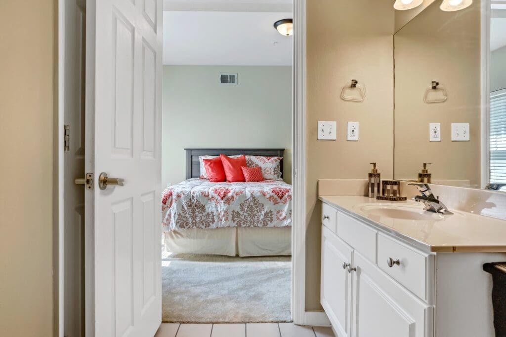 Bright, inviting bathroom leading to a cozy bedroom with elegant bedding and vibrant red accents. Stylish, well-appointed spaces.