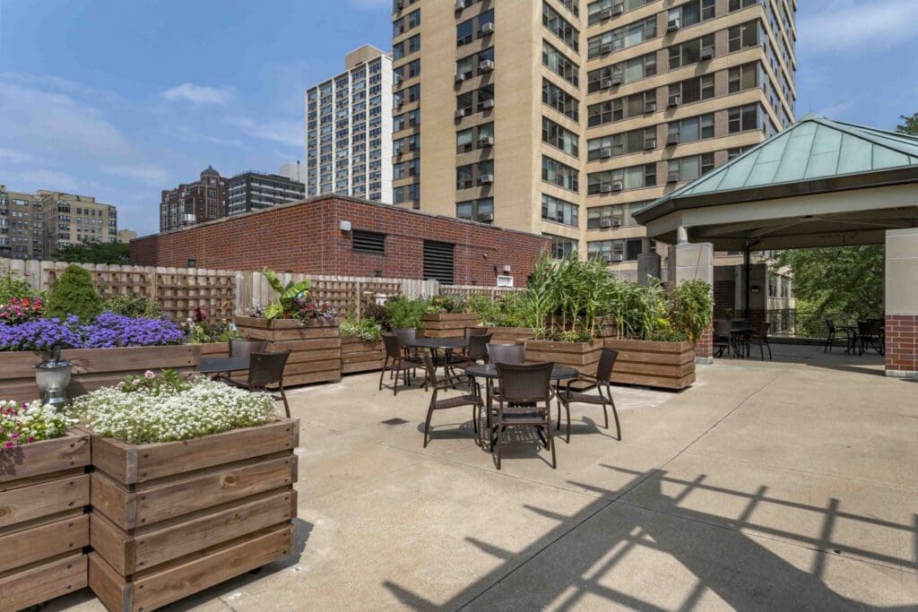 Rooftop garden with wooden planters, colorful flowers, and metal tables and chairs, surrounded by tall urban buildings in the background.